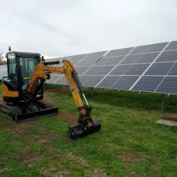 Żółto-czarna minikoparka SANY na gąsienicach, z łyżką w ziemi, na tle rzędu paneli słonecznych, w pochmurny dzień.