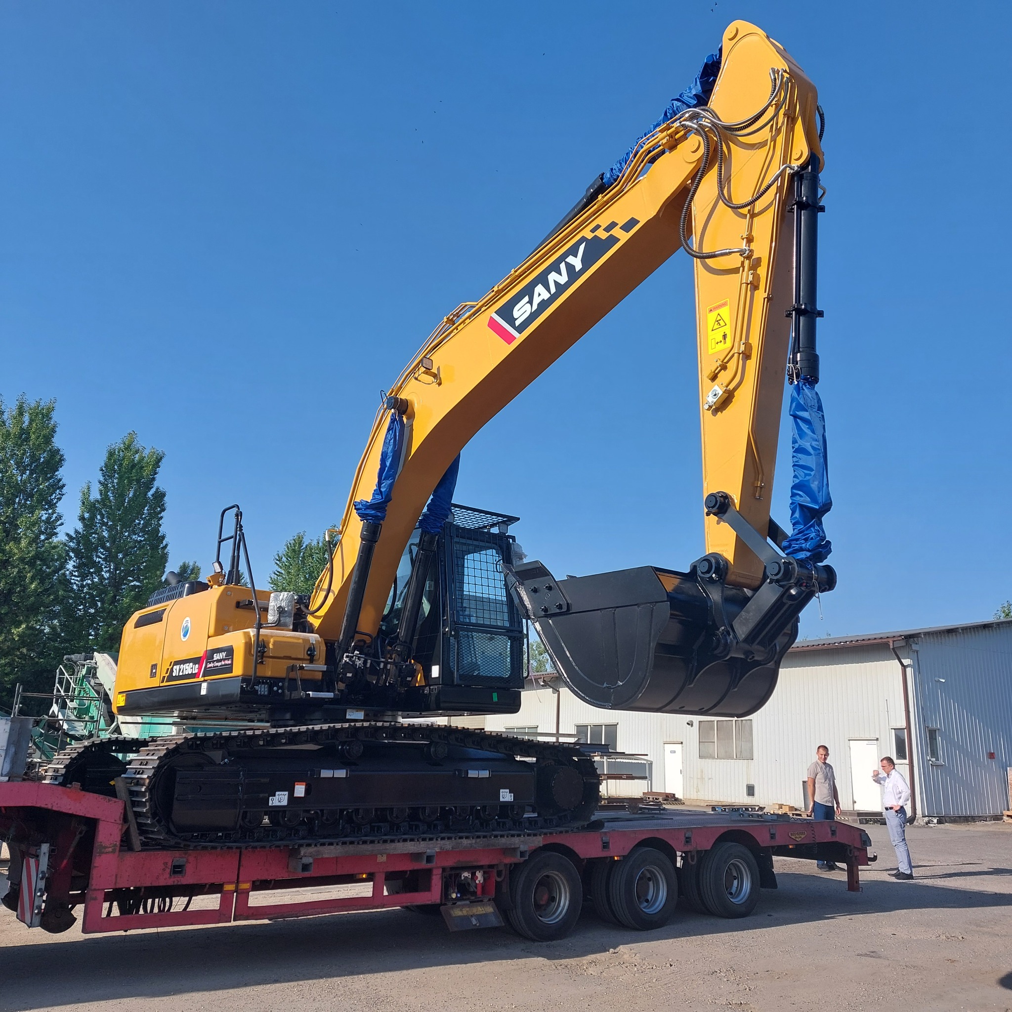 Żółta koparka SANY SY215CLG na czerwonej platformie transportowej, z dwoma mężczyznami obok, na tle błękitnego nieba i budynku przemysłowego.