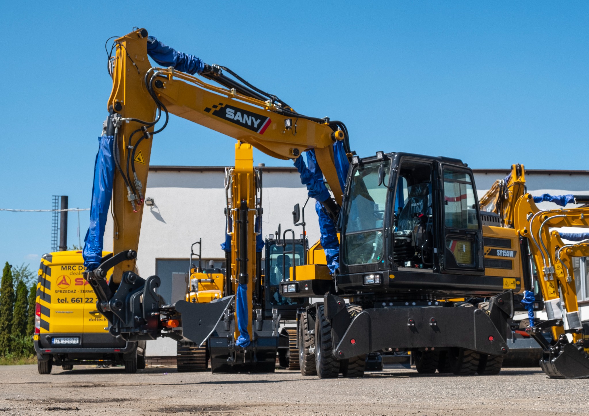 Żółte koparki SANY SY155W z niebieskimi osłonami na siłownikach, ustawione w rzędzie na placu, z żółtym samochodem serwisowym w tle.