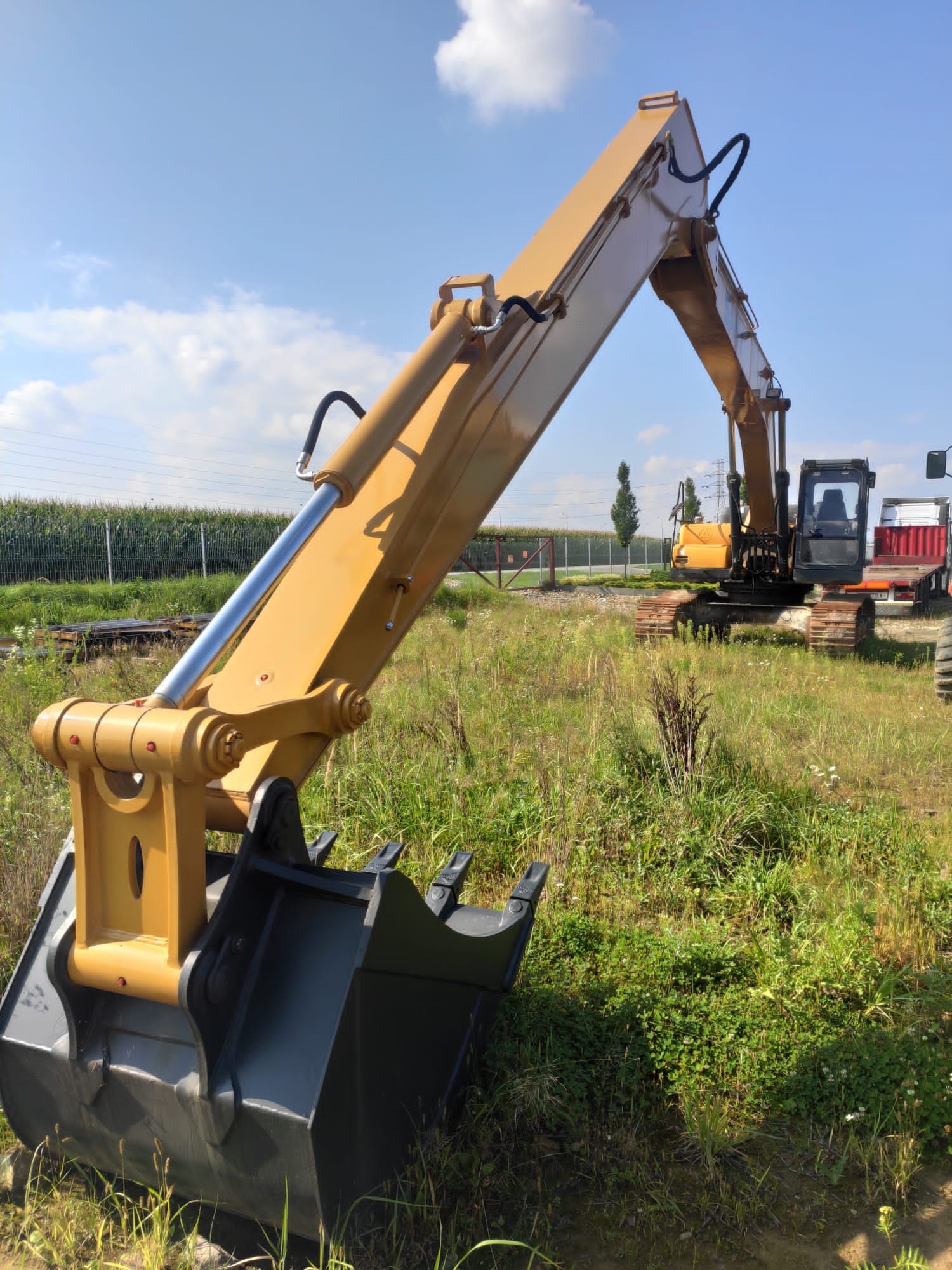 Żółta koparka z czarną łyżką na zewnątrz, widoczna trawa, pole kukurydzy i błękitne niebo z chmurami w tle.