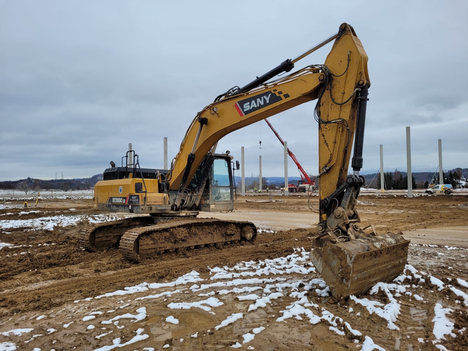Żółta koparka SANY SY265C LC na placu budowy z częściowo stopionym śniegiem, w tle słupy konstrukcyjne i czerwony dźwig.