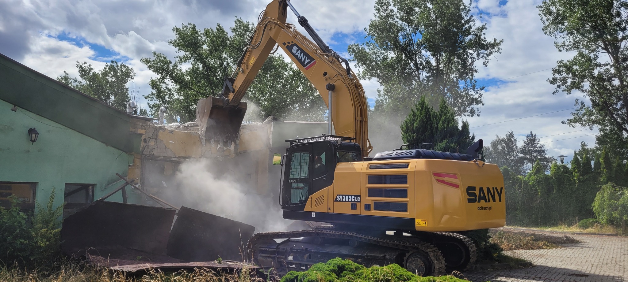 Żółta koparka SANY SY305LC w trakcie rozbiórki budynku, z chmurą pyłu i gruzu w powietrzu, widoczne fragmenty ściany i dachu budynku, tło z drzewami i niebem.