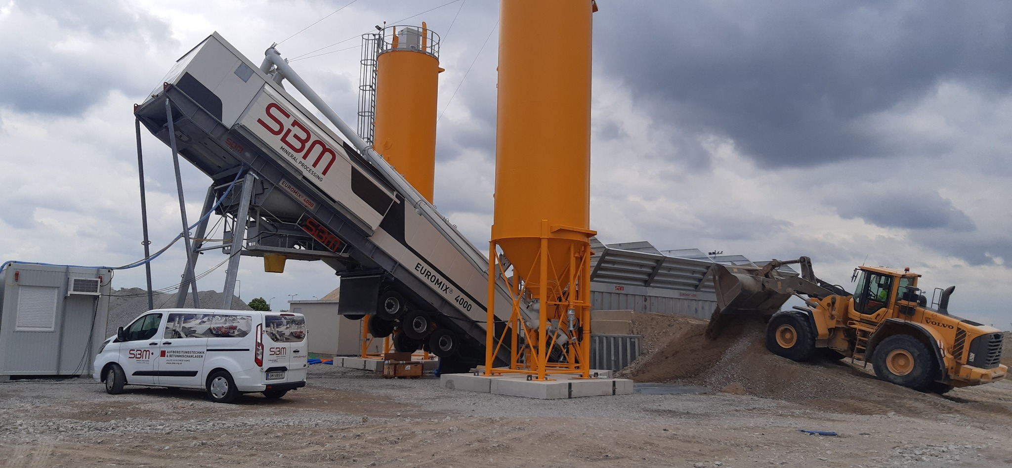 Żółta ładowarka Volvo L150F nabiera piasek na tle mobilnej betoniarni SBM Euromix 4000 z dwoma żółtymi silosami, na placu budowy stoi biały bus firmowy SBM.