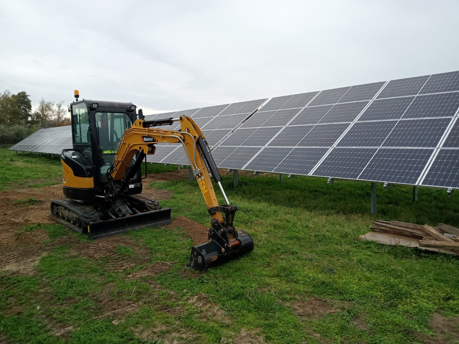 Żółto-czarna minikoparka SANY na gąsienicach, z łyżką w ziemi, na tle rzędu paneli słonecznych, w pochmurny dzień.