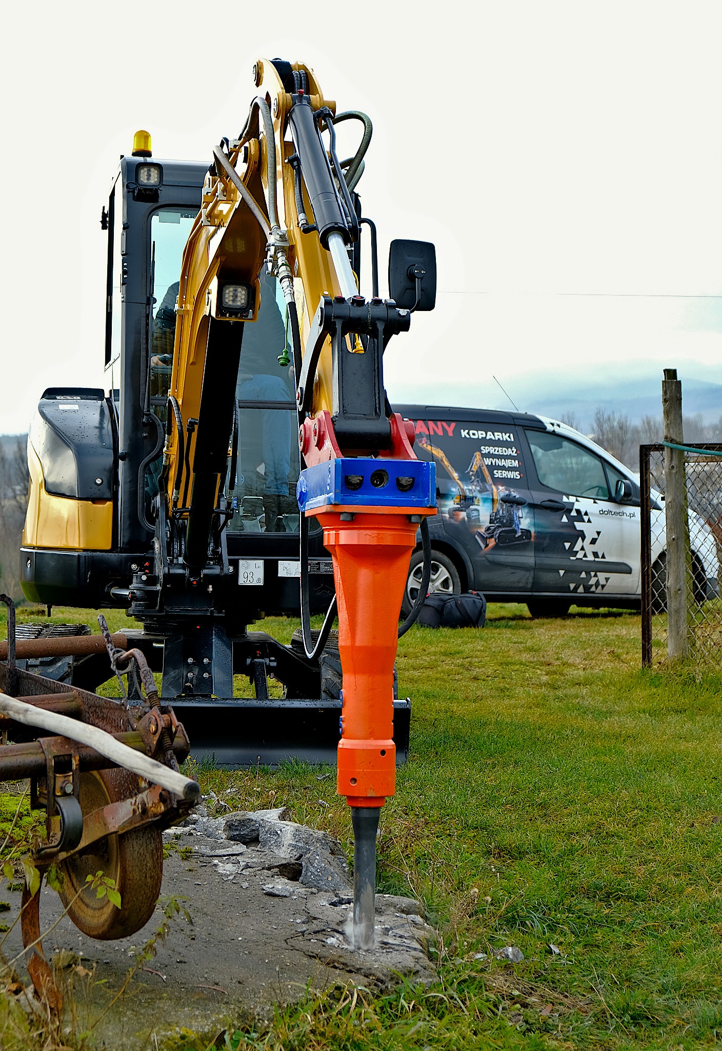 Żółto-czarna minikoparka z zamontowanym młotem hydraulicznym rozbijająca beton, na trawiastym terenie, w tle samochód dostawczy z reklamą firmy budowlanej.
