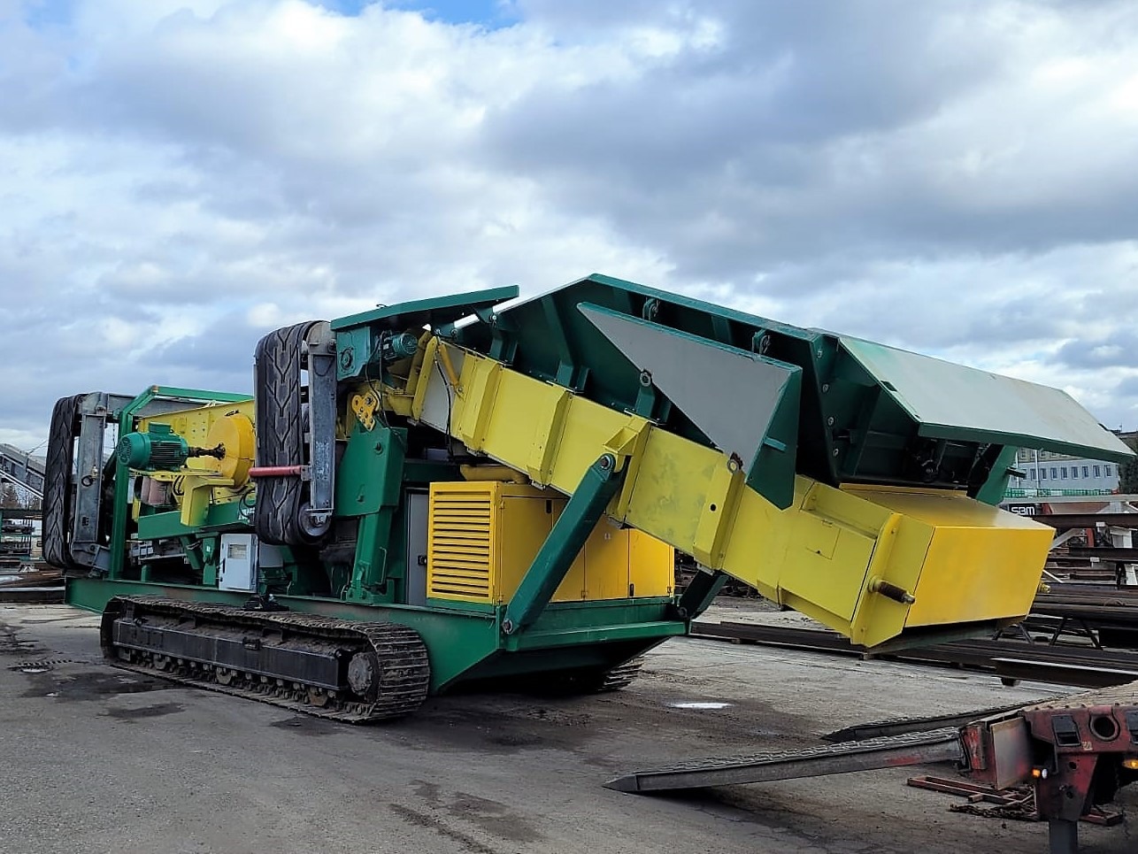 Zielono-żółta maszyna budowlana na gąsienicach z elementami transportującymi materiał, stojąca na placu pod pochmurnym niebem.