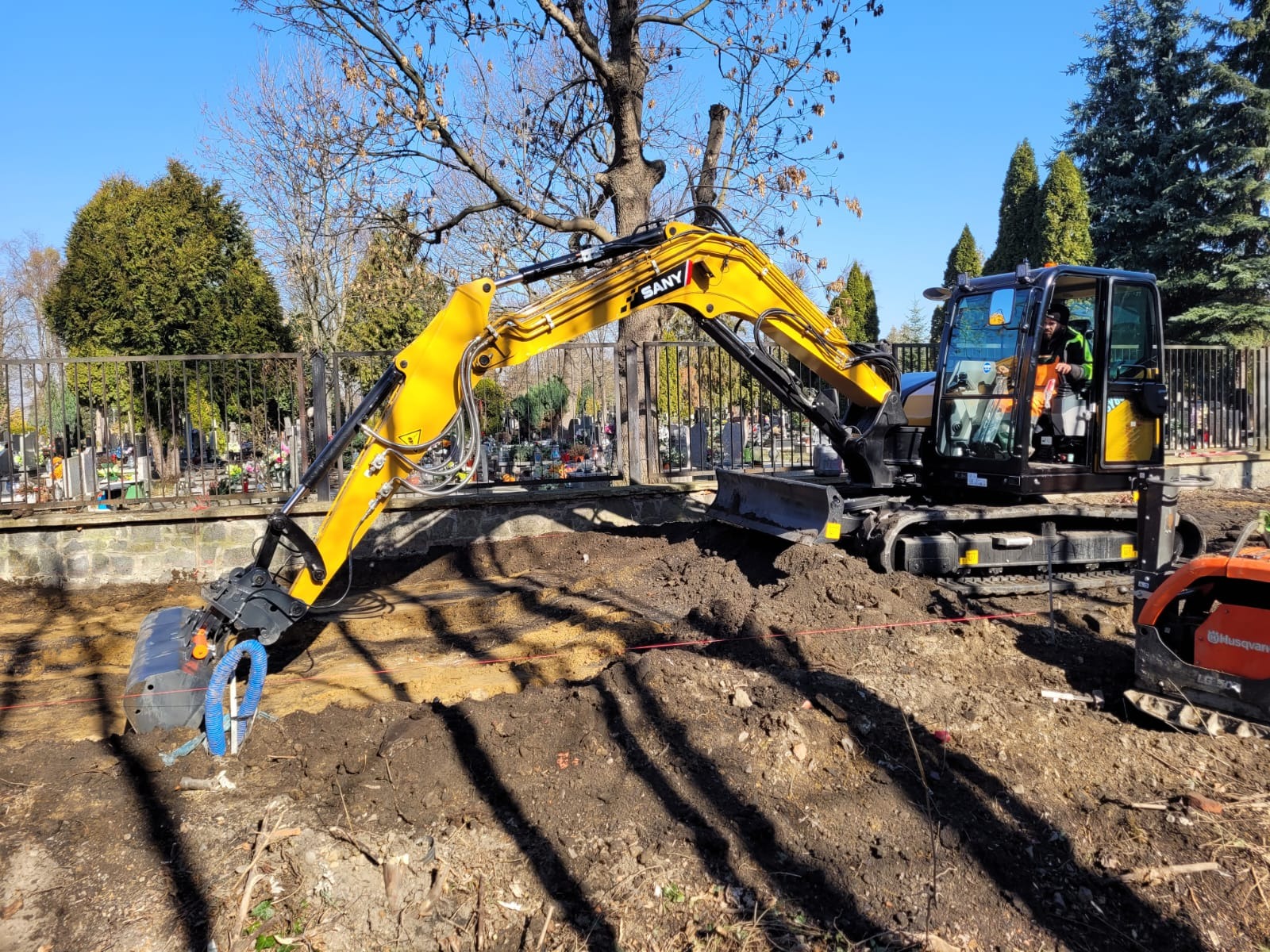 Żółta minikoparka SANY z niebieskimi przewodami hydraulicznymi przy wykopie, w tle cmentarz za metalowym ogrodzeniem.
