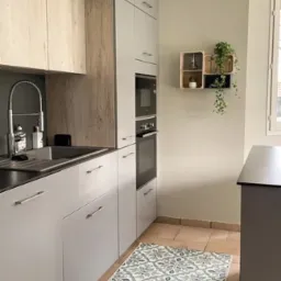 Jasnoszara kuchnia z drewnianymi akcentami, beżową podłogą i dekoracyjnym dywanem w odcieniach zieleni; na ścianie drewniane p&oacute;łki z roślinami doniczkowymi.