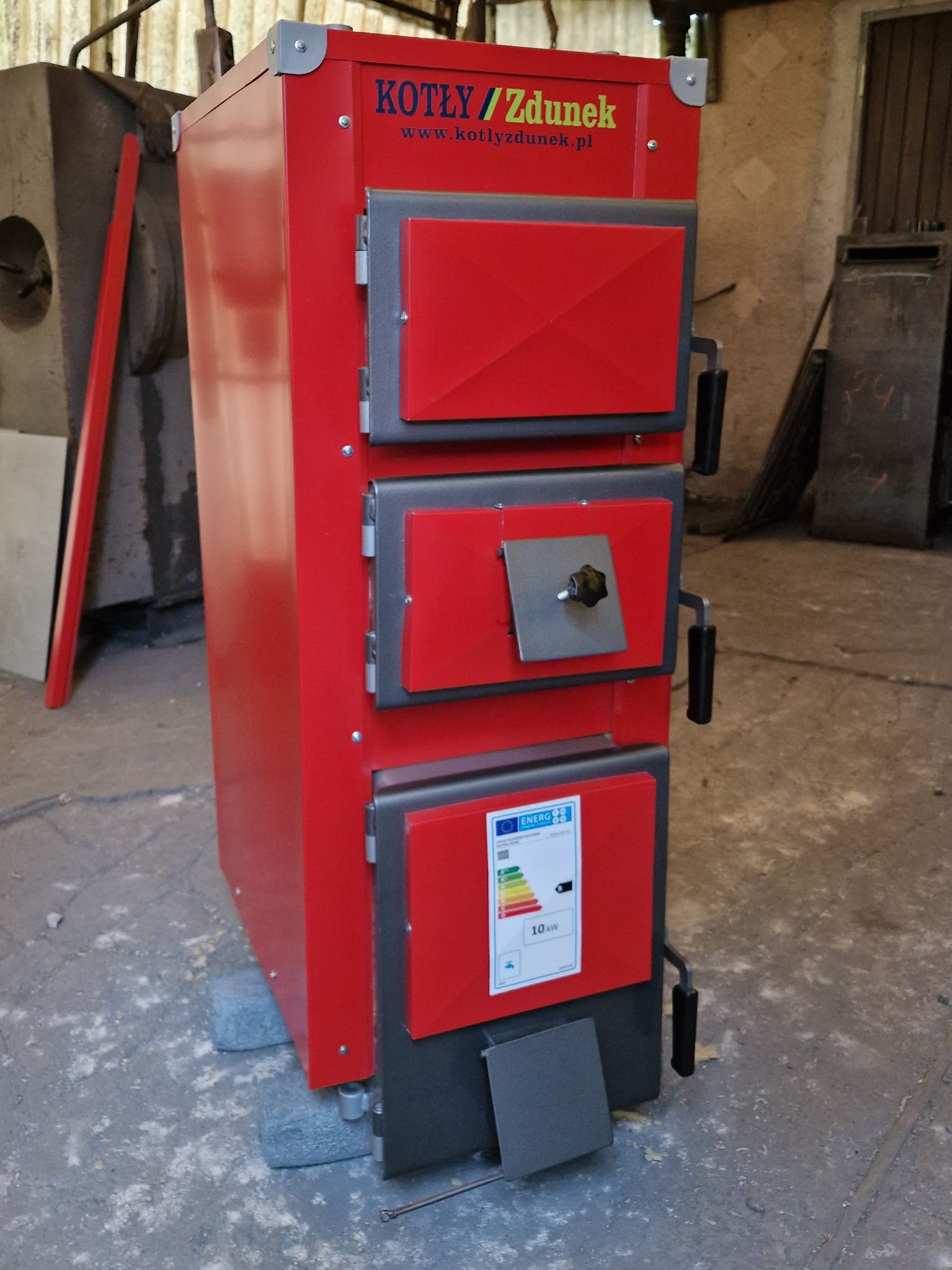 Czerwony, fabrycznie nowy kocioł na paliwo stałe marki Zdunek, z trzema drzwiczkami i etykietą energetyczną, ustawiony na betonowej podłodze w magazynie.