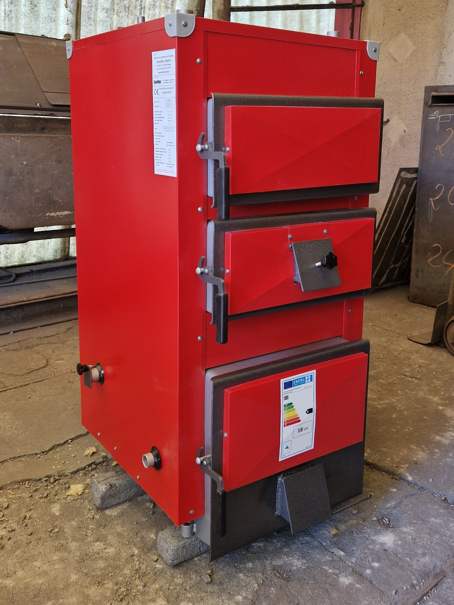 Czerwony, trójdzielny piec na paliwo stałe ustawiony na betonowych bloczkach w surowym wnętrzu, widoczna etykieta energetyczna i uchwyty drzwiczek.