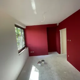 Wnętrze pokoju po częściowym pomalowaniu ścian na intensywny czerwony kolor, widoczne puszki z farbą na szarym, surowym podłożu, biała ściana z oknem po lewej stronie i wystające kable z sufitu.