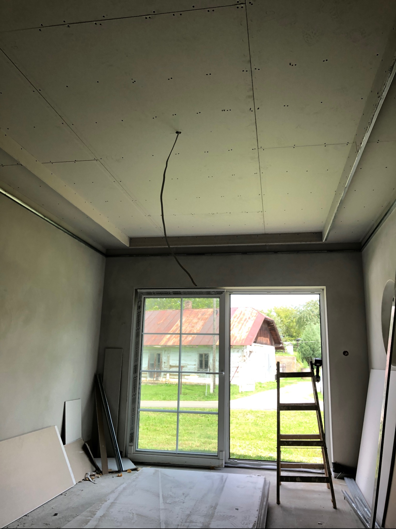 Pokój w trakcie remontu: zamontowany stelaż sufitu podwieszanego z płyt gipsowo-kartonowych, ściany przygotowane pod gładzie, drabina oparta o okno tarasowe.