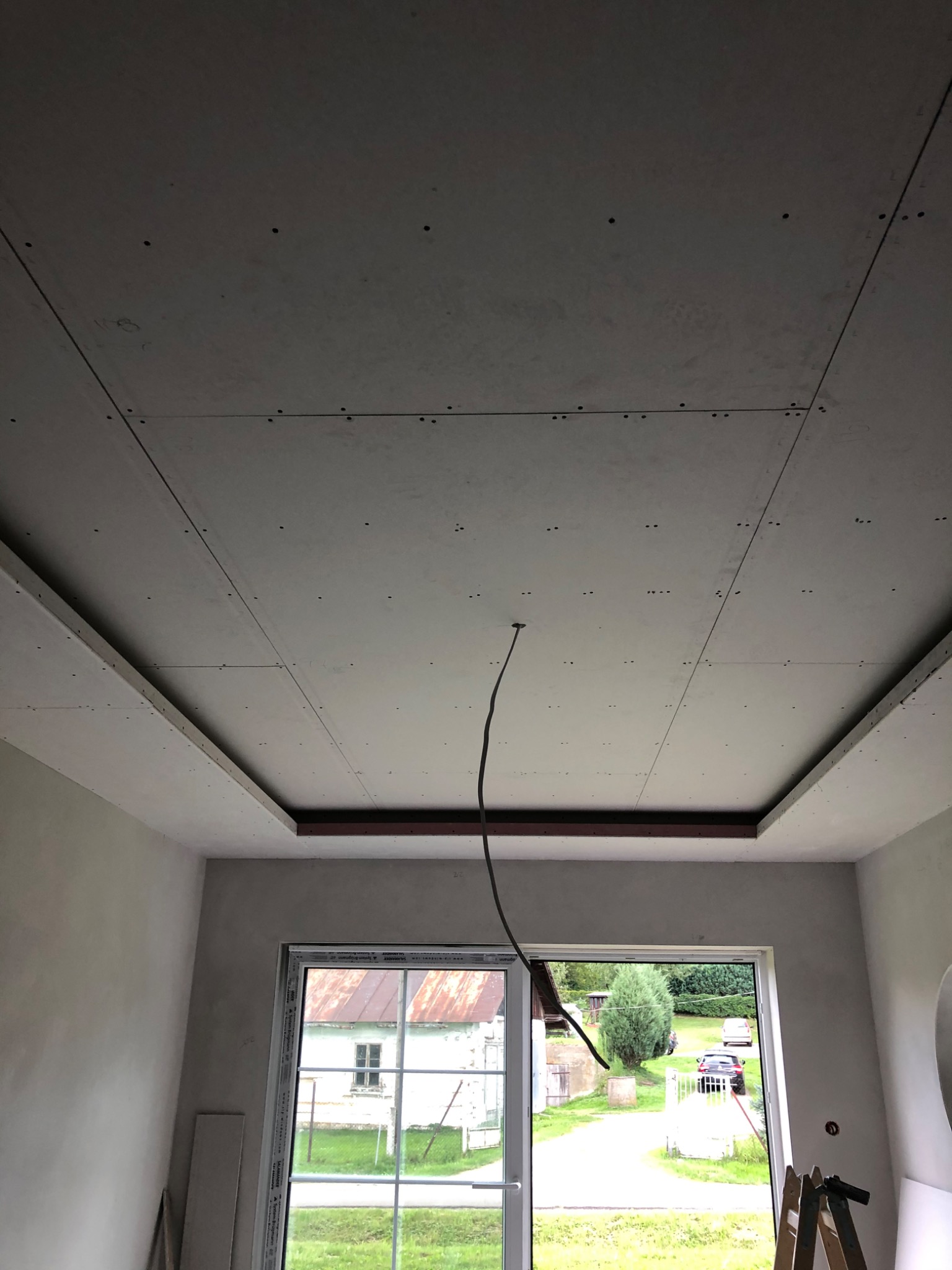 Montaż sufitu podwieszanego z płyt gipsowo-kartonowych z widocznymi łączeniami i wystającym przewodem elektrycznym, okno z widokiem na podwórze.