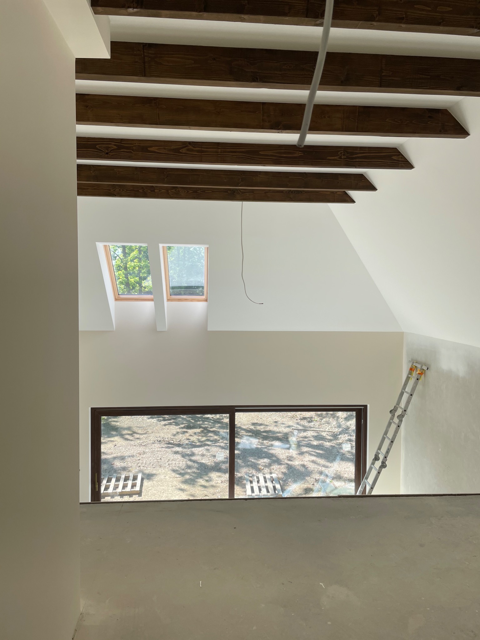 Wnętrze poddasza ze skośnym sufitem, drewnianymi belkami stropowymi, oknami dachowymi i rozsuwanym oknem balkonowym; ściany w trakcie szpachlowania, widoczna drabina aluminiowa.