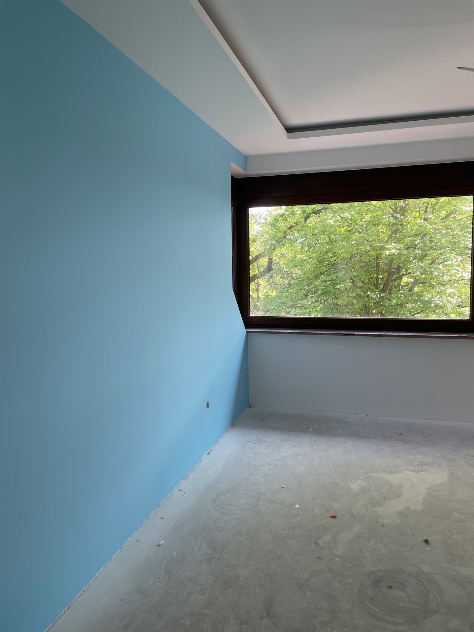 Świeżo pomalowane na błękitno pomieszczenie z brązowym oknem, widok na zieleń drzew za oknem, surowa podłoga przed wykończeniem.