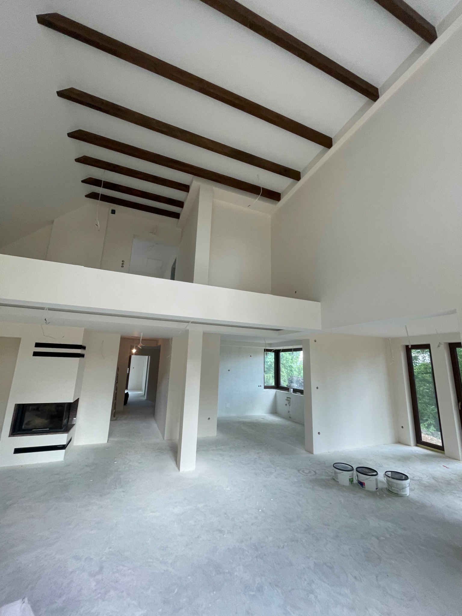 Wnętrze domu w trakcie prac wykończeniowych z widocznymi drewnianymi belkami stropowymi, świeżo pomalowanymi ścianami na biało, nowoczesnym kominkiem i kilkoma puszkami farby na podłodze.