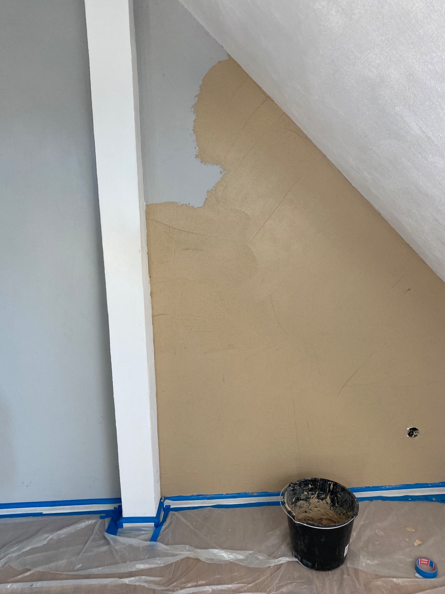 Ściana poddasza w trakcie szpachlowania, widoczny fragment pokryty masą, kubeł z zaprawą na podłodze zabezpieczonej folią i taśmą malarską.