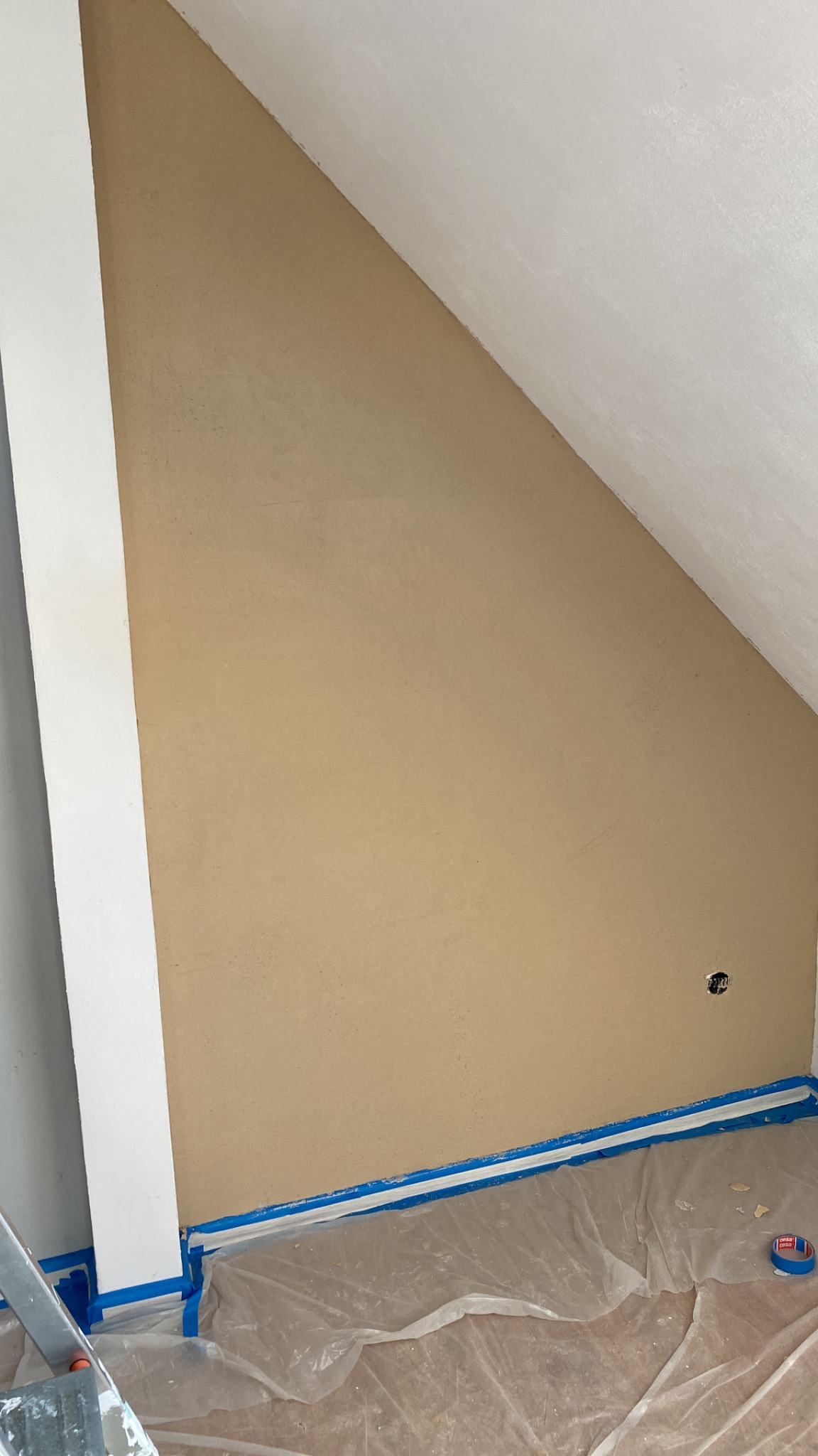 Ściana pod skosem po szpachlowaniu, zabezpieczona taśmą malarską przy podłodze, widoczne gniazdko elektryczne i folia ochronna na podłodze.