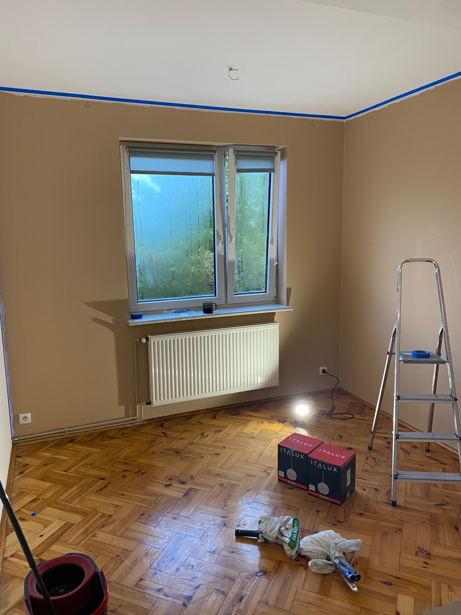 Pokój w trakcie przygotowania do malowania: ściany w kolorze beżowym, zabezpieczone taśmą malarską przy suficie, okno z parą na szybach, grzejnik, drabina malarska, lampa halogenowa i akcesoria...