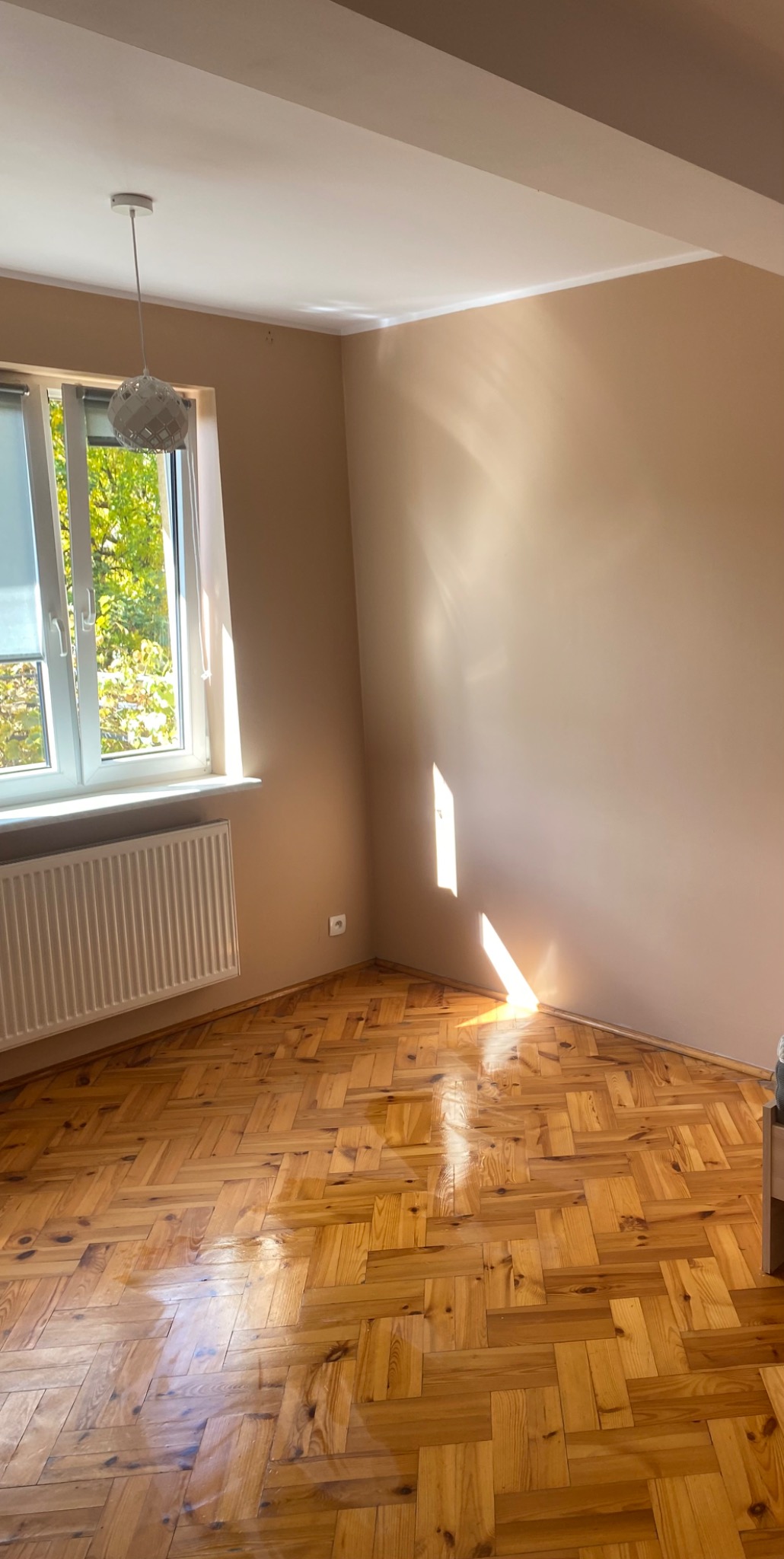 Pomalowane na beżowo wnętrze pokoju z parkietem w jodełkę, białym grzejnikiem pod oknem i designerską lampą wiszącą.
