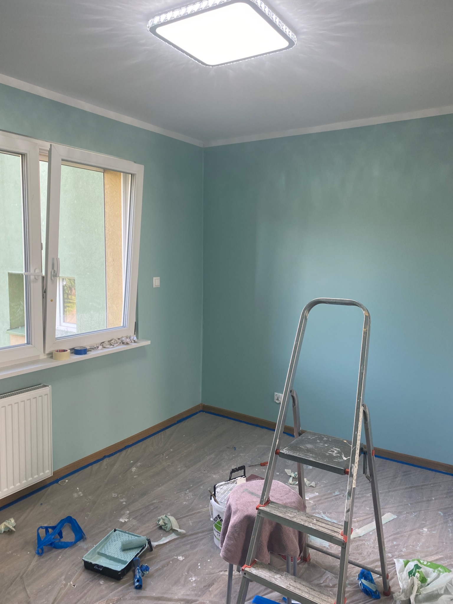 Pokój po malowaniu na pastelowy błękit, drabina oparta o ścianę, podłoga zabezpieczona folią malarską, widoczne narzędzia i akcesoria malarskie.