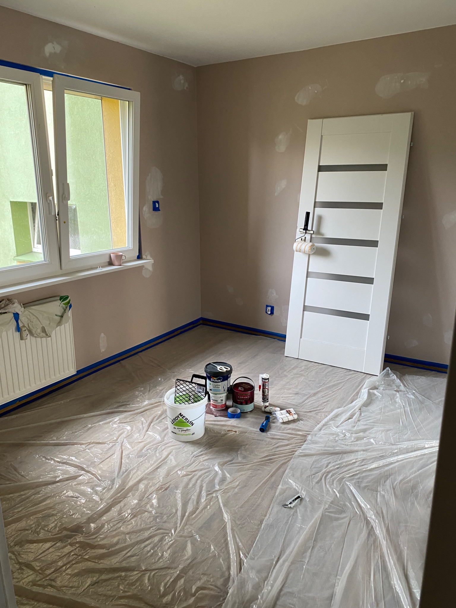 Pokój przygotowany do malowania: zaklejone listwy, folia ochronna na podłodze, ślady szpachli na ścianach, białe drzwi z rolką do malowania, puszki z farbą i narzędzia malarskie.