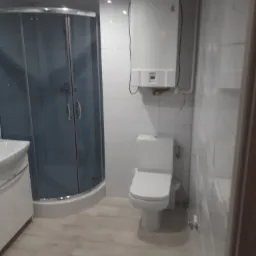 Wykończona łazienka z kabiną prysznicową z szarymi szybami, białą armaturą, grzejnikiem przepływowym i podłogą imitującą drewno.