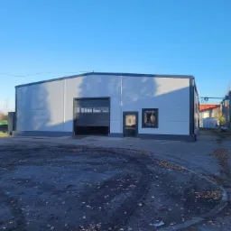 Nowoczesna hala magazynowa z szarej blachy trapezowej, częściowo otwarta brama segmentowa, okno z refleksami i drzwi wejściowe, teren utwardzony z liśćmi.