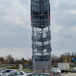 Wysoka, ażurowa konstrukcja stalowa z elementami w kolorach czerwonym i niebieskim, prawdopodobnie pylon reklamowy, na tle parkingu i pochmurnego nieba.