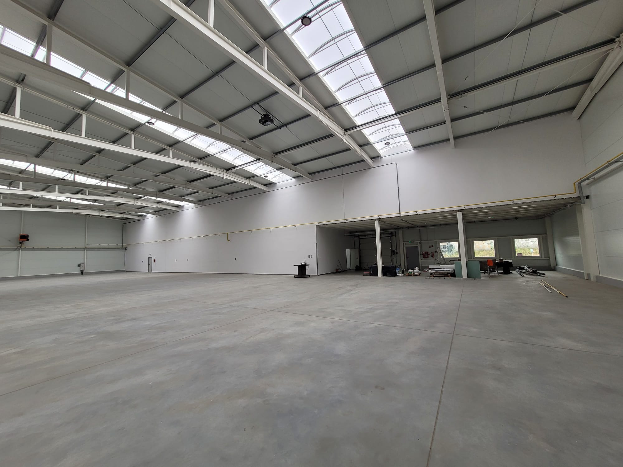 Wnętrze nowo wybudowanej hali z widoczną konstrukcją stalową dachu, oświetleniem górnym i betonową posadzką. Przygotowane do dalszych prac wykończeniowych.