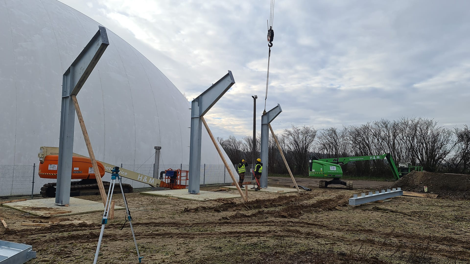 Montaż stalowych słupów pod konstrukcję hali namiotowej, widoczne podnośniki koszowe i pracownicy w żółtych kaskach.