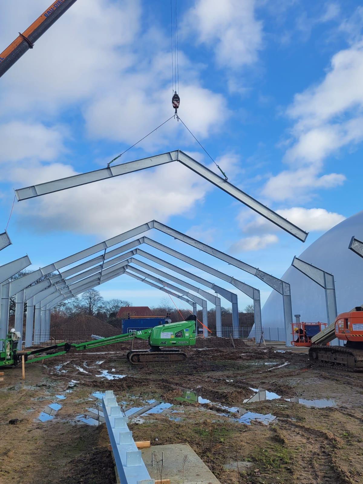 Montaż stalowej konstrukcji hali z użyciem dźwigu na placu budowy, widoczne podnośniki koszowe i błotniste podłoże.
