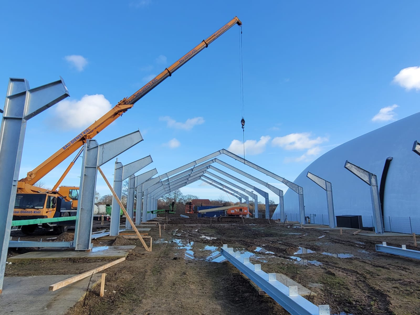 Montaż stalowej konstrukcji hali na błotnistym terenie, widoczny dźwig z napisem 'BAU-DIENST-KIEL', w tle biała kopuła i błękitne niebo.