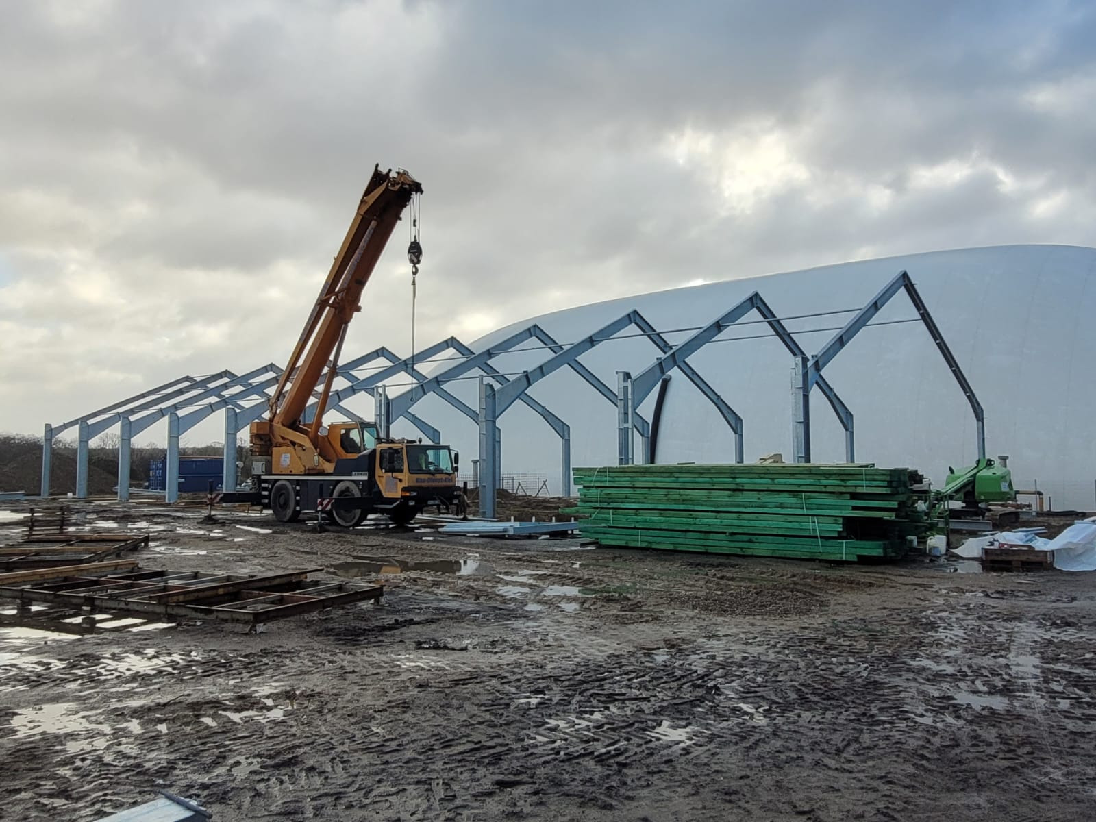 Montaż stalowej konstrukcji hali z użyciem dźwigu na błotnistym terenie, widoczne elementy konstrukcyjne i stos zielonych desek.