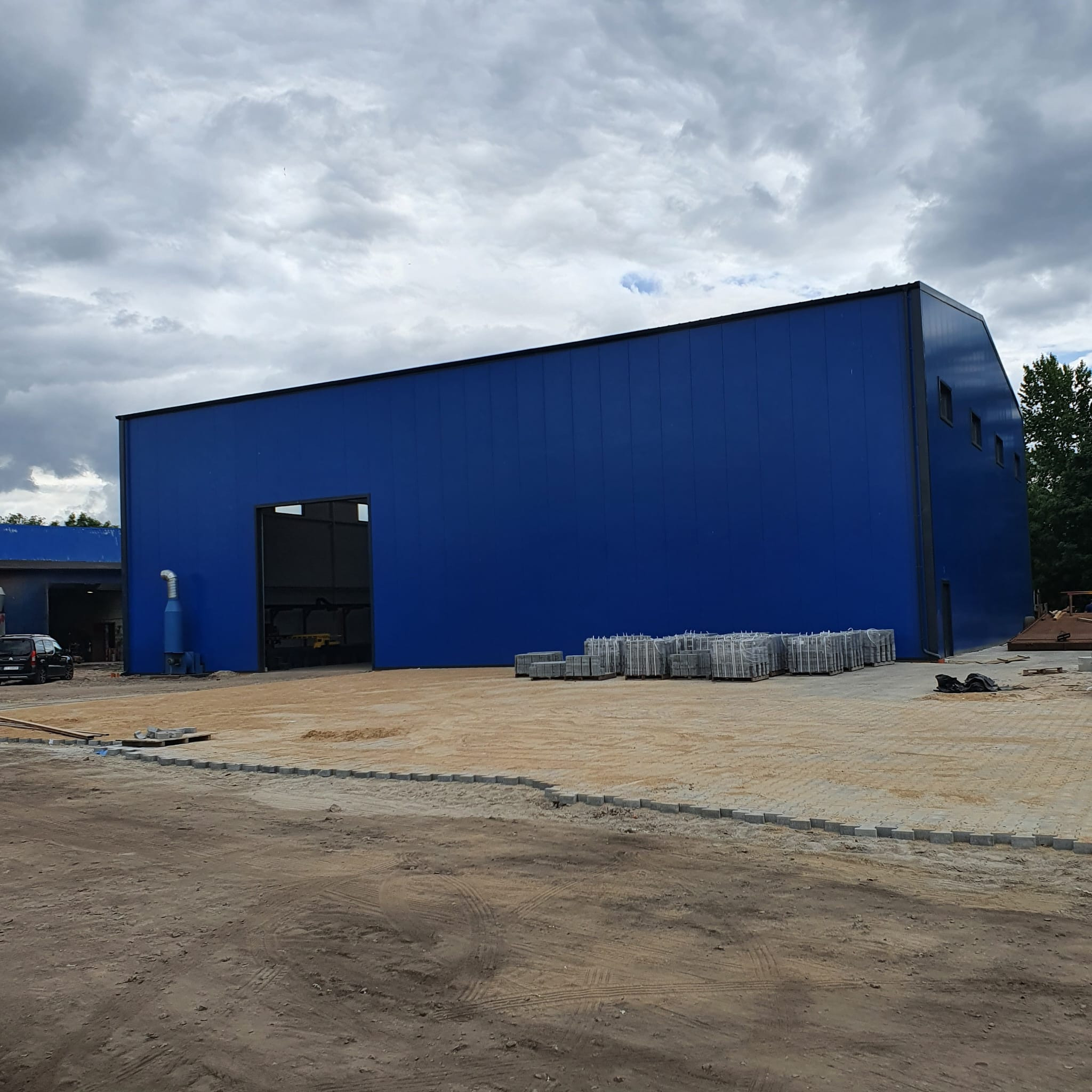 Niebieska hala przemysłowa z płyt warstwowych, widok z zewnątrz, teren przygotowany pod kostkę brukową, palety z kostką obok budynku, pochmurne niebo.