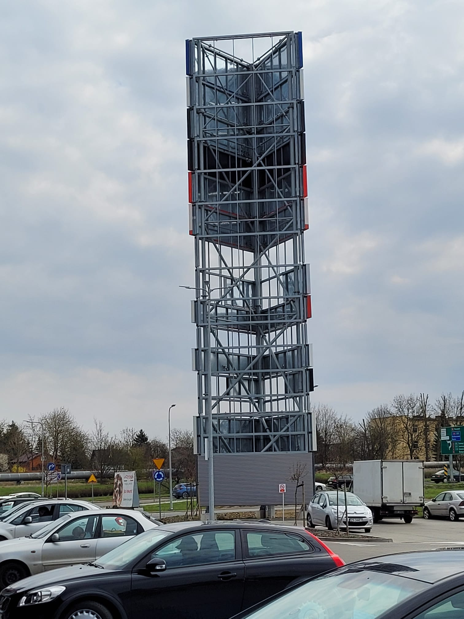 Wysoka, ażurowa konstrukcja stalowa z elementami w kolorach czerwonym i niebieskim, prawdopodobnie pylon reklamowy, na tle parkingu i pochmurnego nieba.