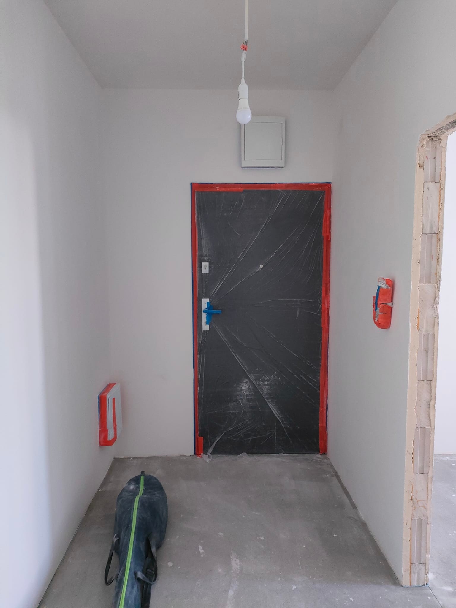 Korytarz w trakcie przygotowań do prac wykończeniowych: ściany pokryte świeżą farbą, drzwi zabezpieczone folią, widoczne taśmy malarskie i torba z narzędziami przy ścianie, surowa żarówka zwisająca...