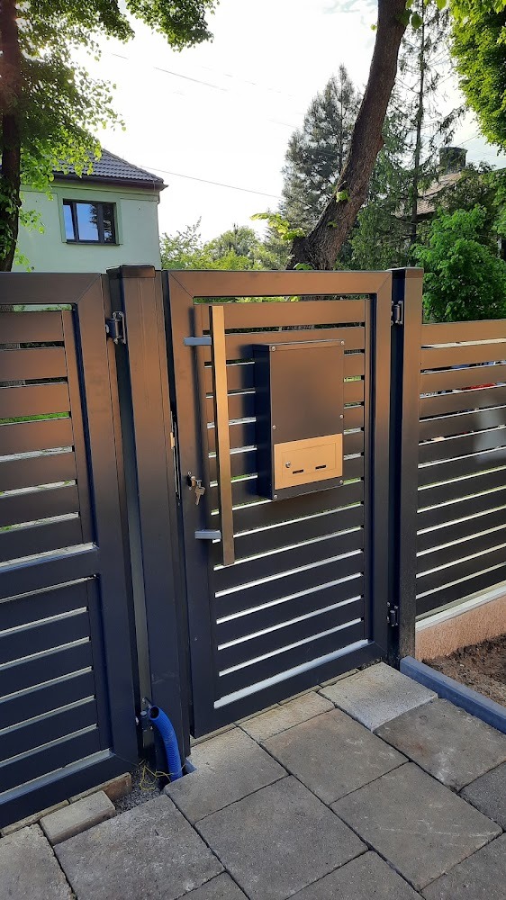 Nowoczesna brama wejściowa z poziomymi panelami, skrzynką na listy i długim uchwytem, widok z zewnątrz na kostkę brukową i dom w tle.