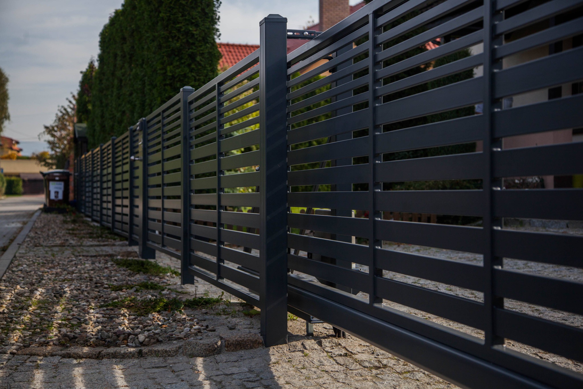 Nowoczesne, poziome, grafitowe przęsła przesuwnej bramy wjazdowej z widocznym mechanizmem jezdnym, na tle brukowanej drogi i zielonych drzew.