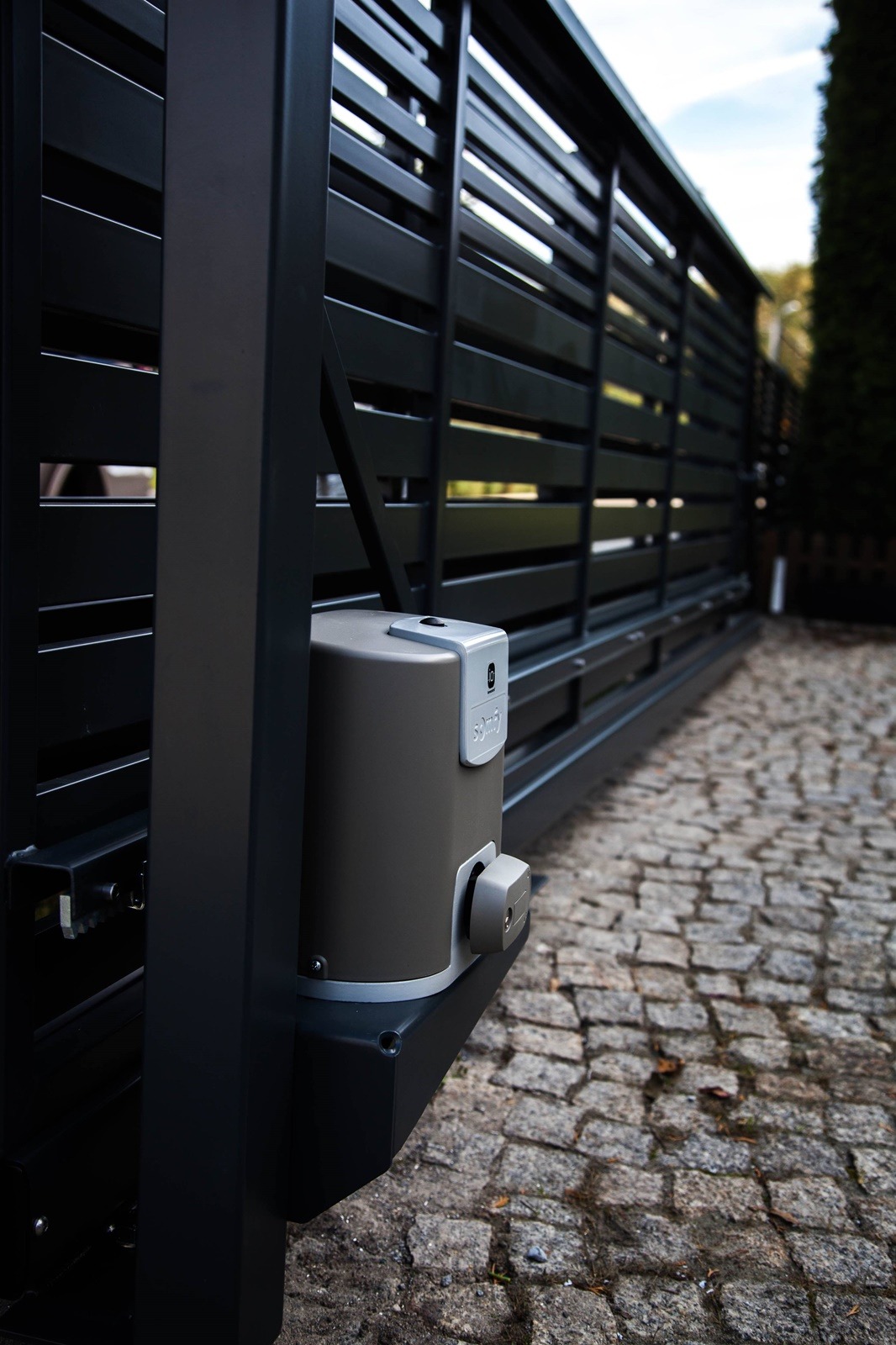 Szara, automatyczna brama przesuwna z czarnymi, poziomymi panelami i napędem Somfy, zamontowana na posesji z kostki brukowej.