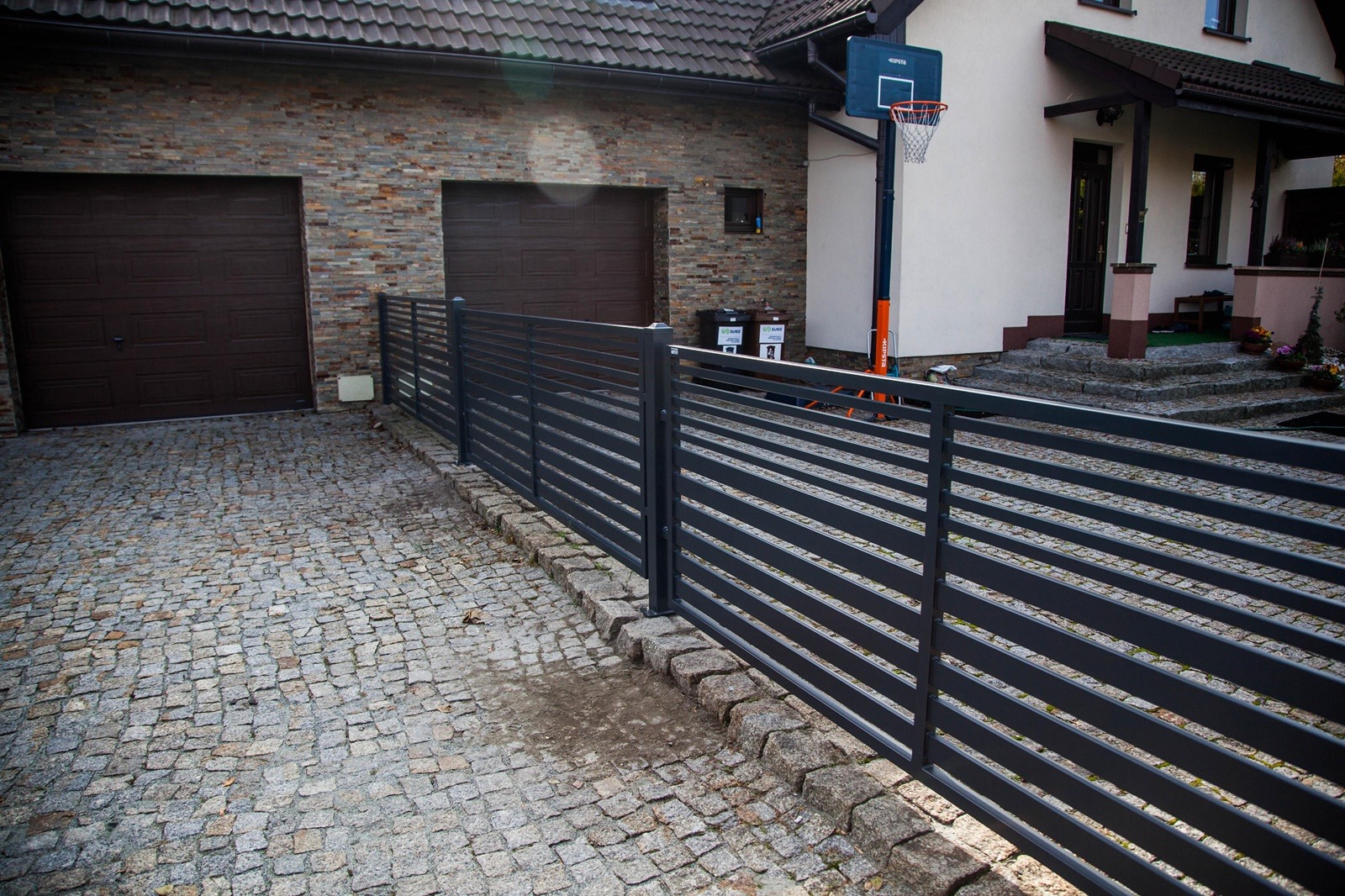 Nowoczesne, poziome, szare przęsła ogrodzeniowe na posesji z kostki brukowej, widok na garaż i dom z elementami kamiennymi.
