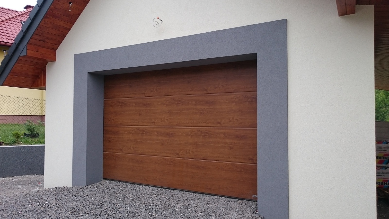 Brązowa, segmentowa brama garażowa z poziomymi panelami imitującymi drewno, osadzona w szarej ramie na tle białej ściany budynku z podsypką z drobnego kamienia.