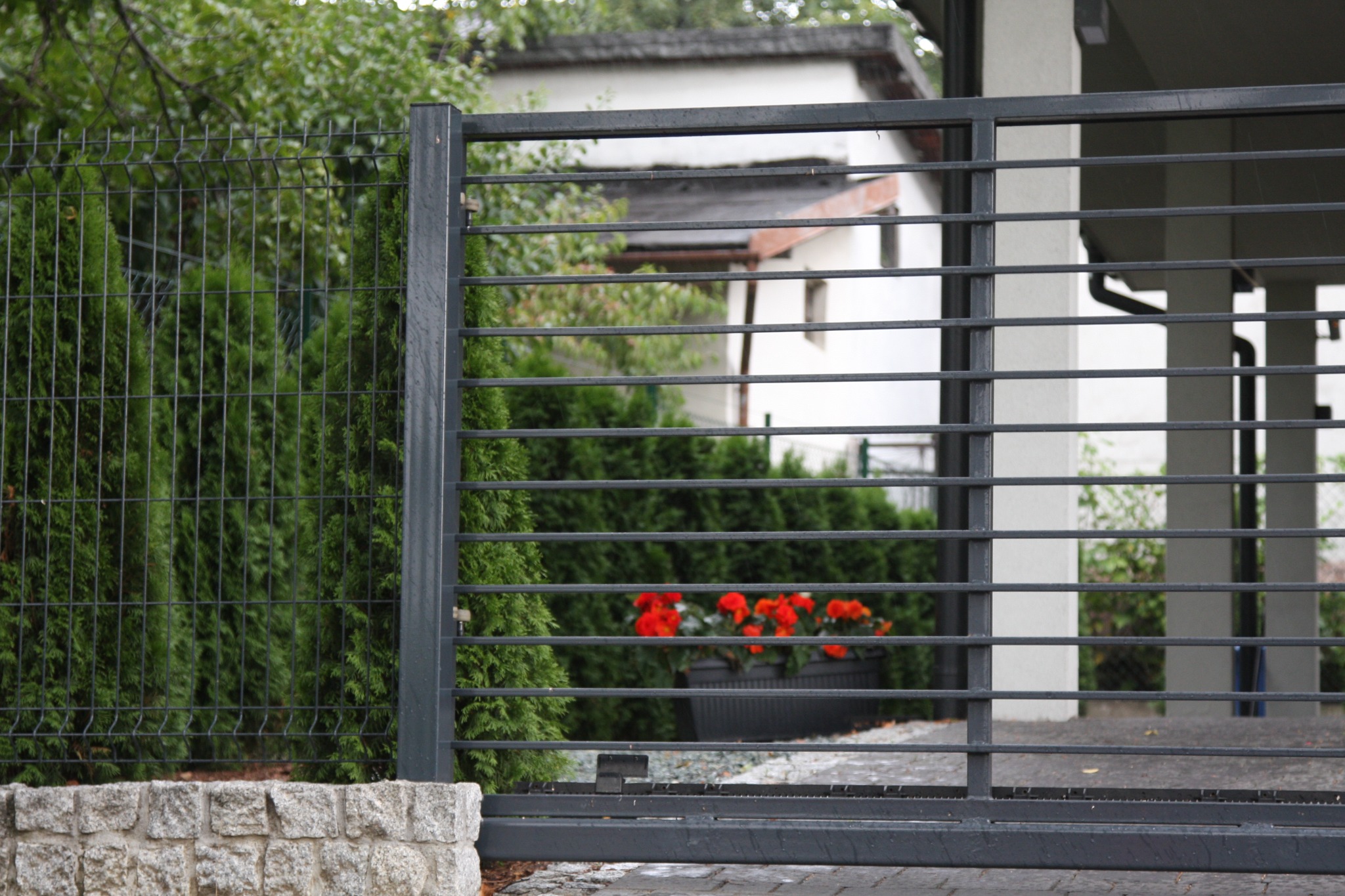 Nowoczesna posesja z bramą przesuwną w kolorze grafitowym, z widocznym fragmentem ogrodzenia panelowego i tujami w tle, ozdobiona donicą z czerwonymi kwiatami.