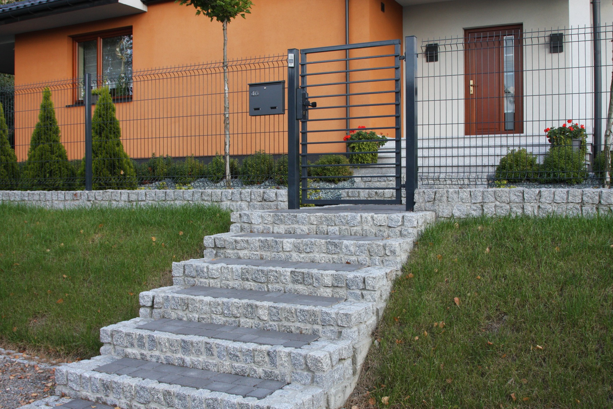 Nowoczesna posesja z ogrodzeniem panelowym, furtką z poziomych profili i kamiennymi schodami wejściowymi, skrzynka na listy przy furtce.
