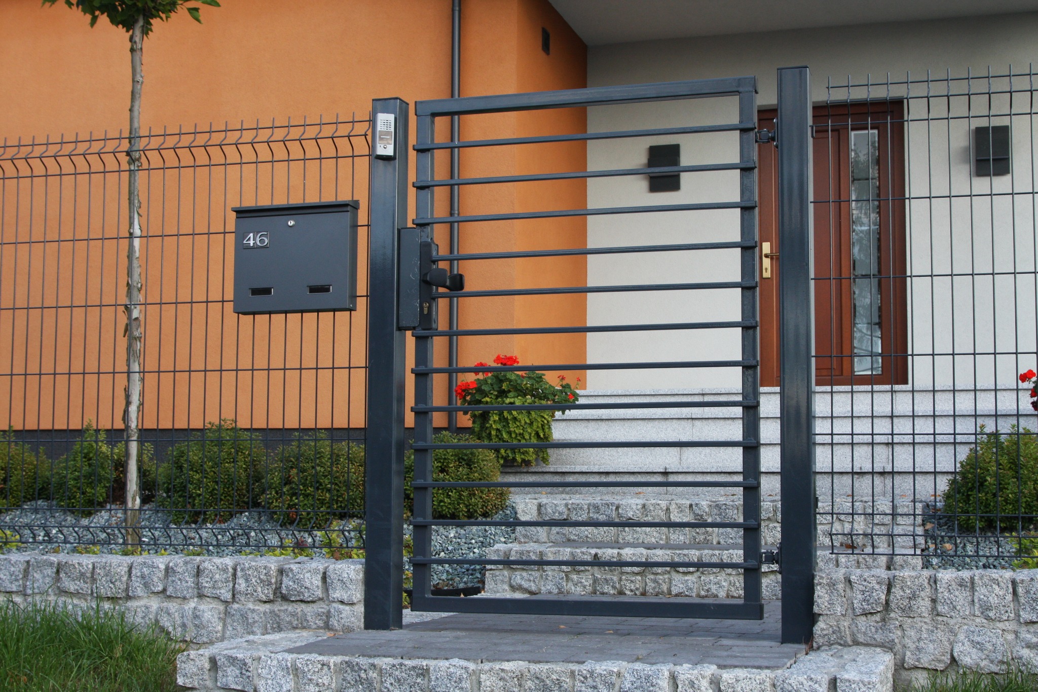 Nowoczesna, grafitowa furtka z poziomymi przęsłami na tle domu z pomarańczową elewacją i skrzynką pocztową numer 46, widoczny fragment ogrodzenia panelowego i kamienne schody.