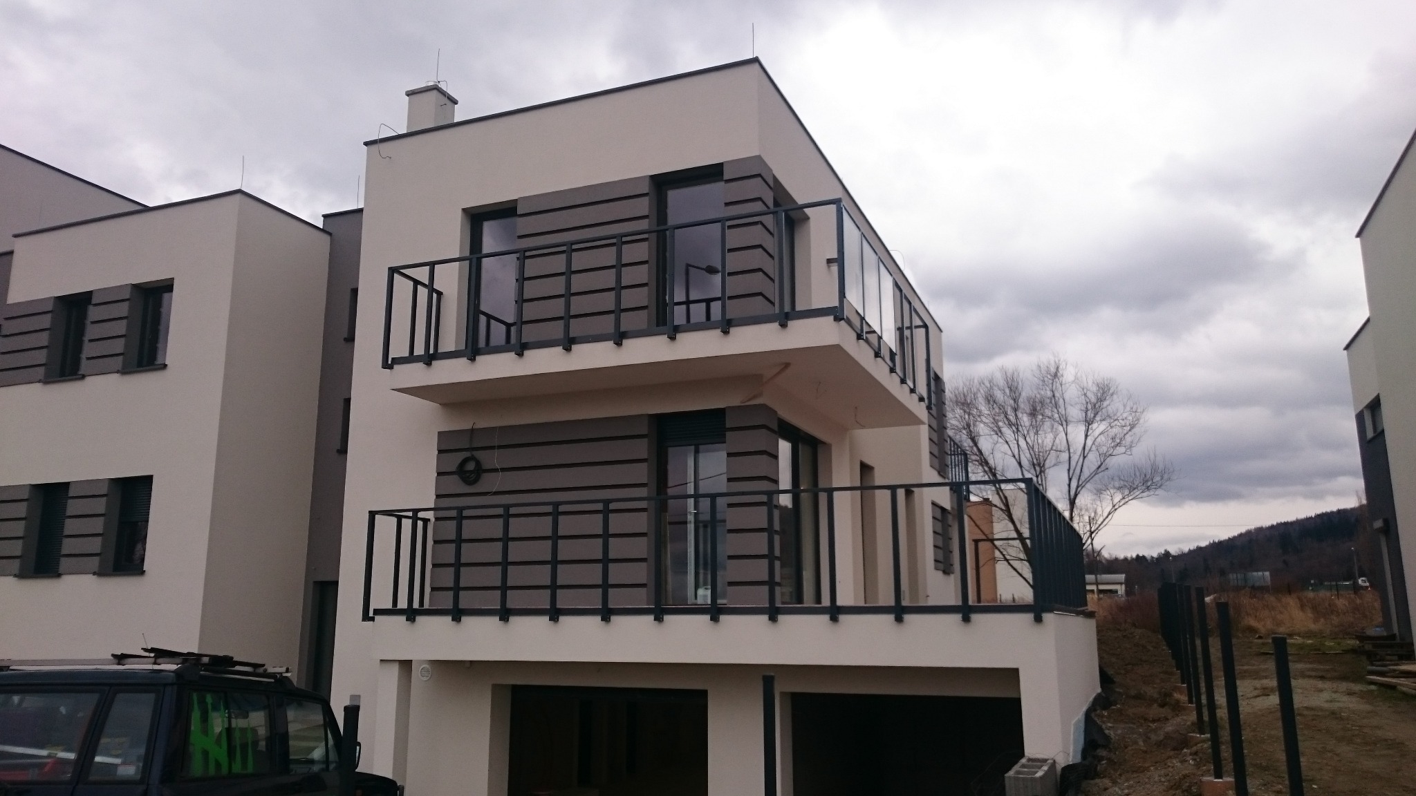 Nowoczesny dom z balkonami zabezpieczonymi metalowymi balustradami i szklanymi panelami, widok na pochmurne niebo i fragment zbocza.