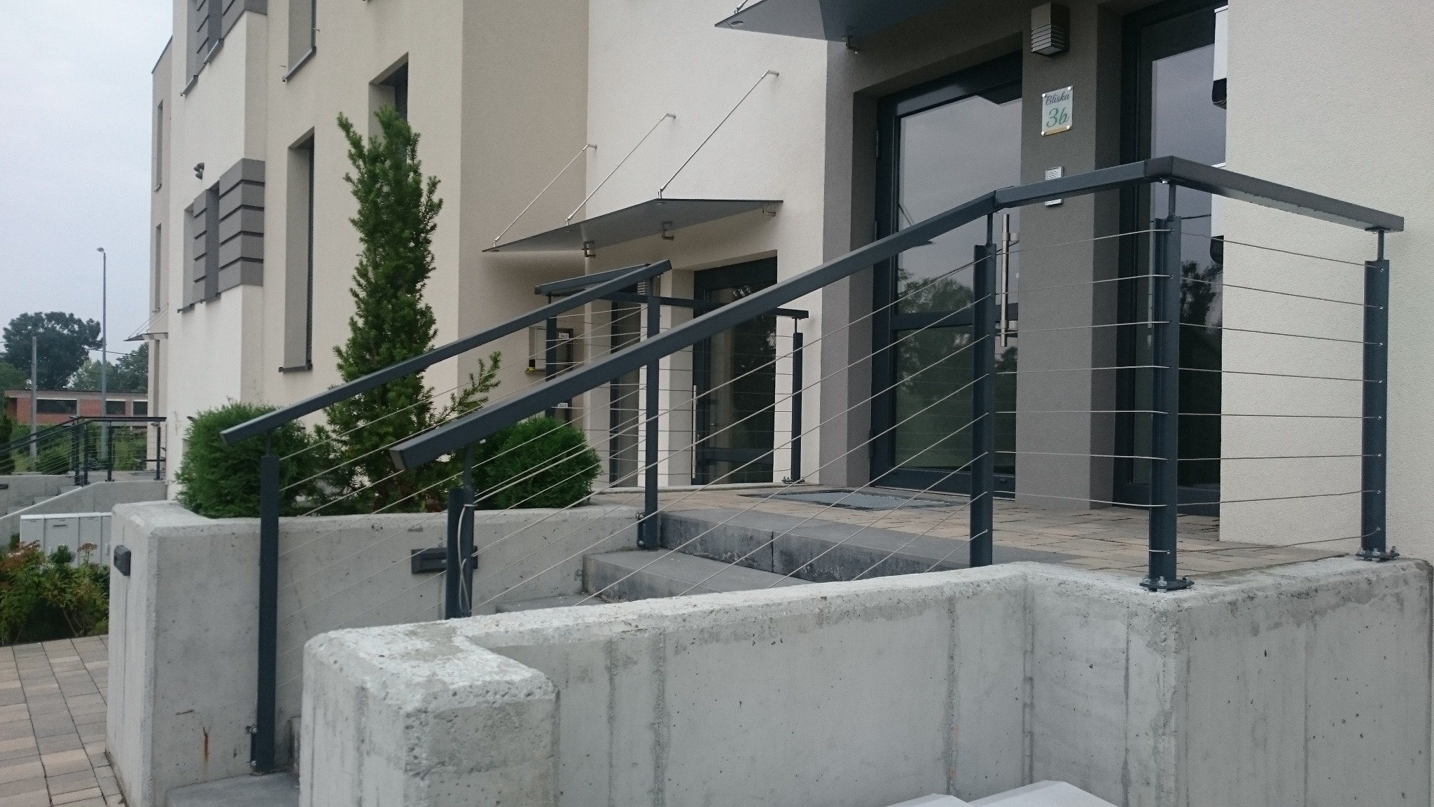 Nowoczesna balustrada schodowa ze stali nierdzewnej z poziomymi linkami, zamontowana na betonowym murku przed wejściem do budynku z szarą elewacją i zadaszeniem.