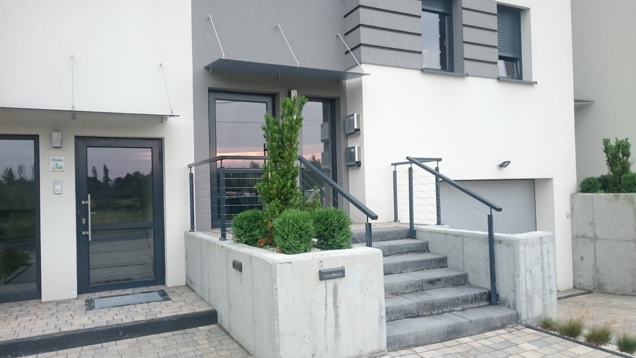 Nowoczesne ogrodzenie metalowe z poziomymi prętami zabezpieczające schody wejściowe do domu z szarymi drzwiami i daszkiem, oraz betonową donicą z roślinami.