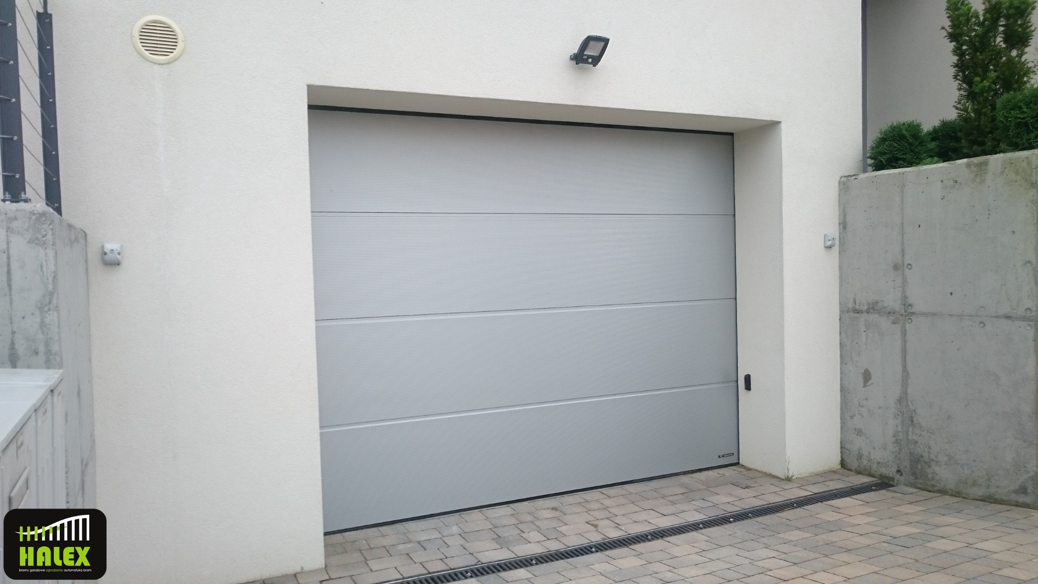 Zamknięta, segmentowa brama garażowa w kolorze jasnoszarym z poziomymi przetłoczeniami, widoczna z zewnątrz budynku, z oświetleniem zewnętrznym nad bramą i logo firmy w lewym dolnym rogu.