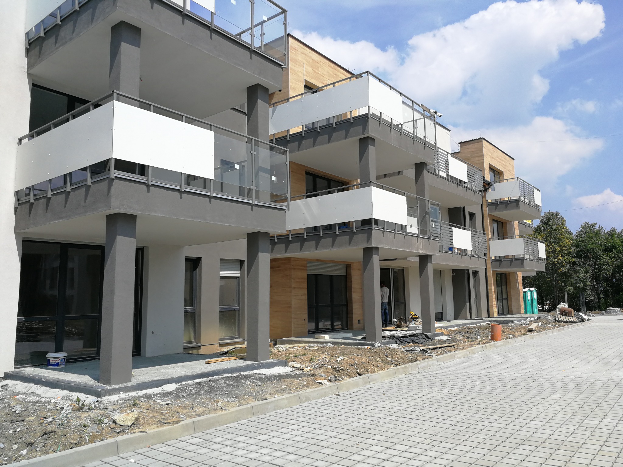 Nowoczesny budynek apartamentowy z balkonami zabezpieczonymi białymi panelami i szklanymi balustradami, widoczny fragment chodnika z kostki brukowej i teren budowy.